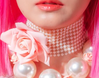 May include: A woman with pink hair wears a pearl necklace and large pearl earrings. She is wearing a white pearl-encrusted choker and a large pink rose.