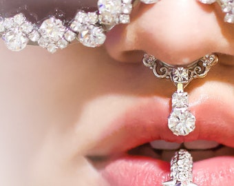 May include: A close-up of a person's face wearing a pair of silver sunglasses with rhinestones, a silver nose ring with a dangling rhinestone, and a silver lip ring with rhinestones.