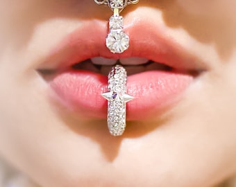 May include: A close-up of a person's mouth with a silver lip ring set with clear rhinestones. The ring has a star-shaped design with pointed edges. The person is also wearing a silver nose ring with a dangling charm set with clear rhinestones.