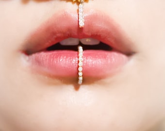 May include: Close-up of a person's lips adorned with a delicate lip ring. The lip ring is a small, gold-colored hoop with a series of tiny, white pearls. A matching nose ring is also visible, adding to the elegant aesthetic.