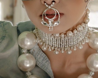 May include: A silver bunny mask with long ears. The person is wearing a pearl necklace and a jeweled nose ring. The background is a light green wall and a window.