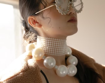 May include: A woman wearing a brown fuzzy jacket, a black lace top, and a pearl necklace. She is wearing large, round, silver-rimmed sunglasses with a sparkly design.