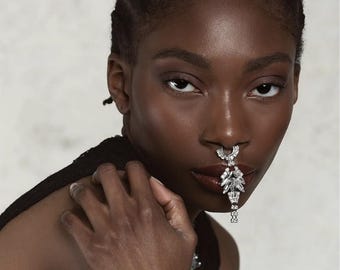 May include: A woman with short, braided hair is wearing a large, silver nose ring with rhinestones. The nose ring is shaped like a teardrop and hangs down to her chin.