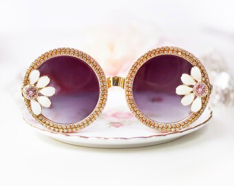 May include: Round sunglasses with a gold frame and rhinestones. The sunglasses have a gradient purple lens and a white flower design on each side.