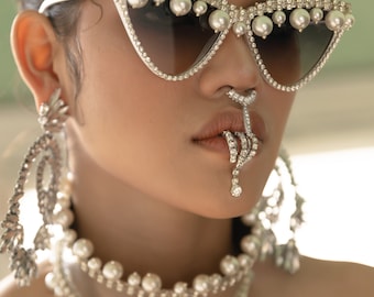 May include: Close-up of a woman wearing elaborate jewelry. White cat-eye sunglasses are adorned with pearls and rhinestones. A jeweled nose ring and lip jewelry are also visible, along with a pearl necklace and large earrings.