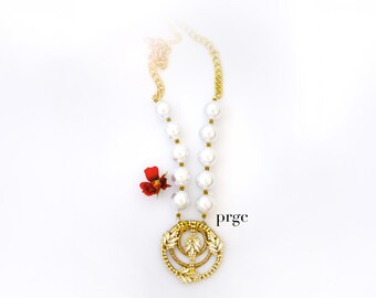 May include: A gold-toned necklace with large white pearls and a decorative pendant. The pendant features a circular design with leaf accents. A small red flower is placed near the necklace. The text "prgc" is visible.