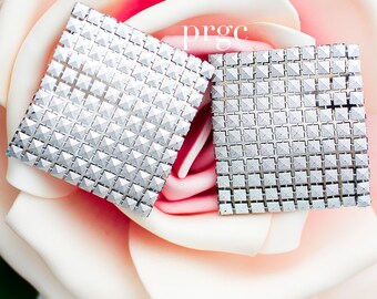May include: Two silver square-shaped cufflinks with a geometric pattern of small pyramids. The cufflinks are displayed on a pink and white rose.