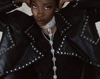 May include: A woman wearing a black leather jacket with silver studs and a silver crystal necklace. The jacket has a collar and a zipper closure.