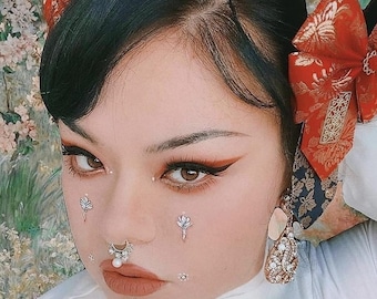 May include: A woman with dark hair and red bows in her hair is wearing a white shirt. She has a nose ring and several facial piercings. Her makeup is dramatic with black eyeliner and red eyeshadow.