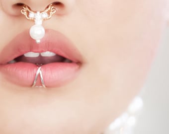May include: Close-up of facial jewelry. A gold-colored nose ring with a decorative design and a dangling pearl. A silver lip ring is also visible. The jewelry is set against a light skin tone.
