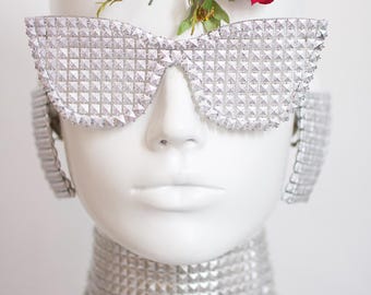 May include: A mannequin head adorned with silver studded sunglasses, ear cuffs, and a choker. A red rose and green foliage are placed on top of the head. The accessories have a geometric pattern.