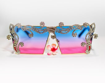 May include: A pair of blue and pink gradient sunglasses with a gold frame and rhinestones. The sunglasses have a decorative design on the frame.