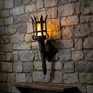 May include: A wall-mounted torch with a black metal frame and a simulated flame. The torch is mounted on a stone wall with a wooden shelf below. The torch has a dark brown handle and a black mounting bracket.