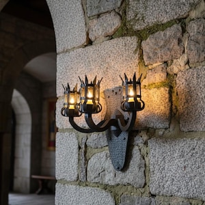 May include: A black wrought iron wall sconce with three candle-style lights. The sconce is mounted on a rough stone wall. The lights are enclosed in decorative cages, and the overall design has a medieval aesthetic.
