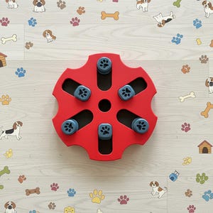 May include: A red dog puzzle toy with six blue compartments, each featuring a paw print design. The toy is on a light wood-grain surface decorated with dog and bone illustrations, and paw prints.
