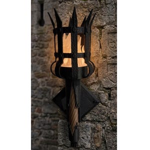 May include: A black metal wall-mounted torch with a decorative cage design and a diamond-shaped base. The torch is mounted on a weathered stone wall, and the flame inside is glowing. The torch has a medieval or gothic aesthetic.