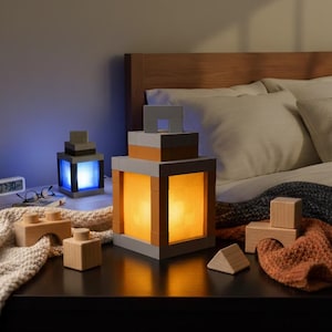 May include: Two illuminated block-style lamps with a warm glow sit on a nightstand. The lamps are constructed of gray and orange blocks. Wooden toy blocks and a digital clock are also on the nightstand. The lamps are next to a bed with white pillows.