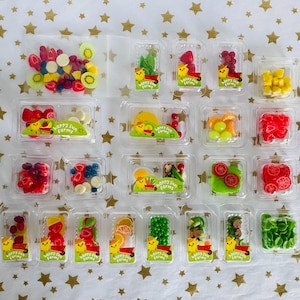 May include: A collection of miniature fruit and vegetable-themed candies in clear plastic containers. The candies include strawberries, raspberries, kiwi, and more. Each container has a "Happy Sun Farms" sticker. The background is a gold star pattern.