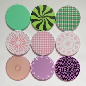 May include: Nine round, colorful phone grips with various designs. The designs include solid green, a green and black swirl, a green and red plaid, a pink floral pattern, a pink gingham, a pink floral plate, a clear orange, a purple with a white border, and a black and purple checkerboard.