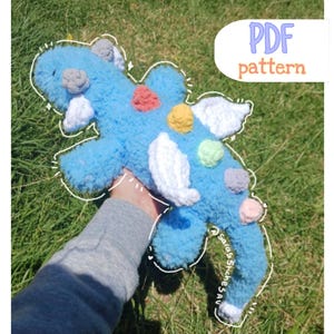 May include: A handmade blue plush toy with white wings and colourful accents. The toy has a fluffy texture and is decorated with various coloured yarn balls. The image includes the text "PDF pattern".