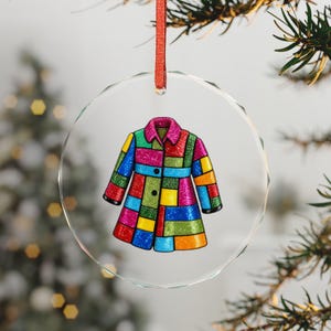 Könnte beinhalten: Ein durchsichtiger, runder Glasanhänger mit einer Illustration eines bunten Patchwork-Mantels mit glitzernden Texturen in Pink, Blau, Gelb, Grün und Orange, der an einem dünnen roten Band zum Aufhängen am Weihnachtsbaum befestigt ist.