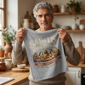 May include: A light gray kitchen towel with the text "SAINTS at the KITCHEN TABLE" in a vintage style. The towel features an illustration of people at a table and the words "PRAY * CHAT * SNACK".