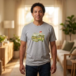 May include: A gray t-shirt featuring a colorful graphic of a golf course with the word "Masters" above it. The graphic is in the shape of the state of Georgia. The person is wearing blue jeans.
