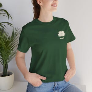 Puede incluir: Una camiseta verde oscuro con un pequeño gráfico en el pecho. El gráfico es una imagen estilizada de un edificio con el texto "You Bet!" impreso encima. La camiseta está hecha de un material suave.