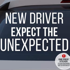 Könnte beinhalten: Weißer Aufkleber mit dem Text "NEW DRIVER EXPECT THE UNEXPECTED" in fetter, serifenloser Schrift. Ein kleiner, runder Aufkleber unten rechts lautet "ONE PIECE STICKER CLEAR BACKING NOT LETTER DECALS". Der Aufkleber ist auf einer Autoscheibe angebracht.