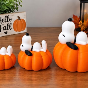 May include: Three white Snoopy figurines with black ears, each lying on top of a bright orange pumpkin. The figurines are arranged in a row from smallest to largest, with each Snoopy figure resting in a relaxed pose on the smooth, rounded pumpkin surface.