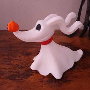May include: A white ghost dog figurine with long, flowing ears, a black eye, and a red collar. The ghost dog has a small, bright orange pumpkin nose. The figure is sculpted in a smooth, matte material and is posed in a dynamic, forward-leaning stance.