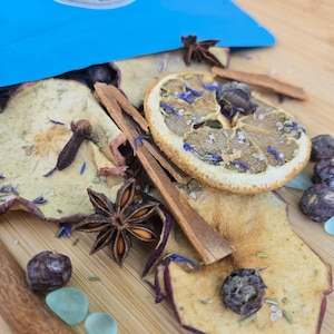 May include: A mixture of dried apple slices, star anise, cinnamon sticks, cloves, dark berries, and small light blue stones. A bright blue pouch with a circular label reading Samhain is positioned at the top of the dried botanical blend.