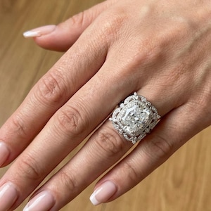 Könnte beinhalten: Ein aufwendiger Verlobungsring mit einem großen, rechteckigen Mittelstein. Der Ring hat einen Halo aus kleineren Steinen und aufwendige Details am Band, gefasst in Silber. Der Ring wird an einer Hand mit gepflegten Nägeln gezeigt.