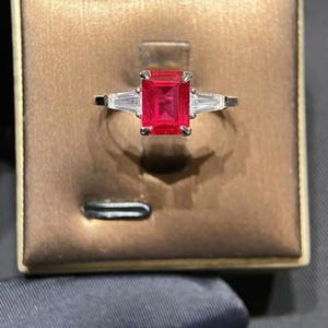 May include: A silver ring with a large, rectangular, bright red gemstone as the focal point. Two smaller, rectangular, clear gemstones flank the main stone. The ring is set in a brown jewelry box.