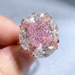 以下が含まれることがあります： ローズゴールドのリングのクローズアップ。大きなピンク色のクッションカットダイヤモンドが特徴です。中央の石は、白いペアシェイプダイヤモンドと小さなピンク色のアクセントストーンで囲まれています。