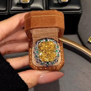 May include: A large, yellow gemstone ring with a square cut, surrounded by smaller blue and green stones. The ring is presented in a brown velvet ring box. Other rings are visible in the background.