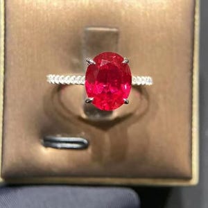 Könnte beinhalten: Ein silberner Ring mit einem großen, oval geschliffenen, leuchtend roten Edelstein als Mittelpunkt. Das Band ist mit kleinen, funkelnden Steinen besetzt. Der Ring wird in einer braunen Schmuckschatulle präsentiert, was ein luxuriöses Gefühl erzeugt.