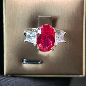 May include: A silver ring featuring a large, oval-cut, vibrant red gemstone as the centerpiece, flanked by two smaller, rectangular, clear gemstones. The ring is set against a brown background.