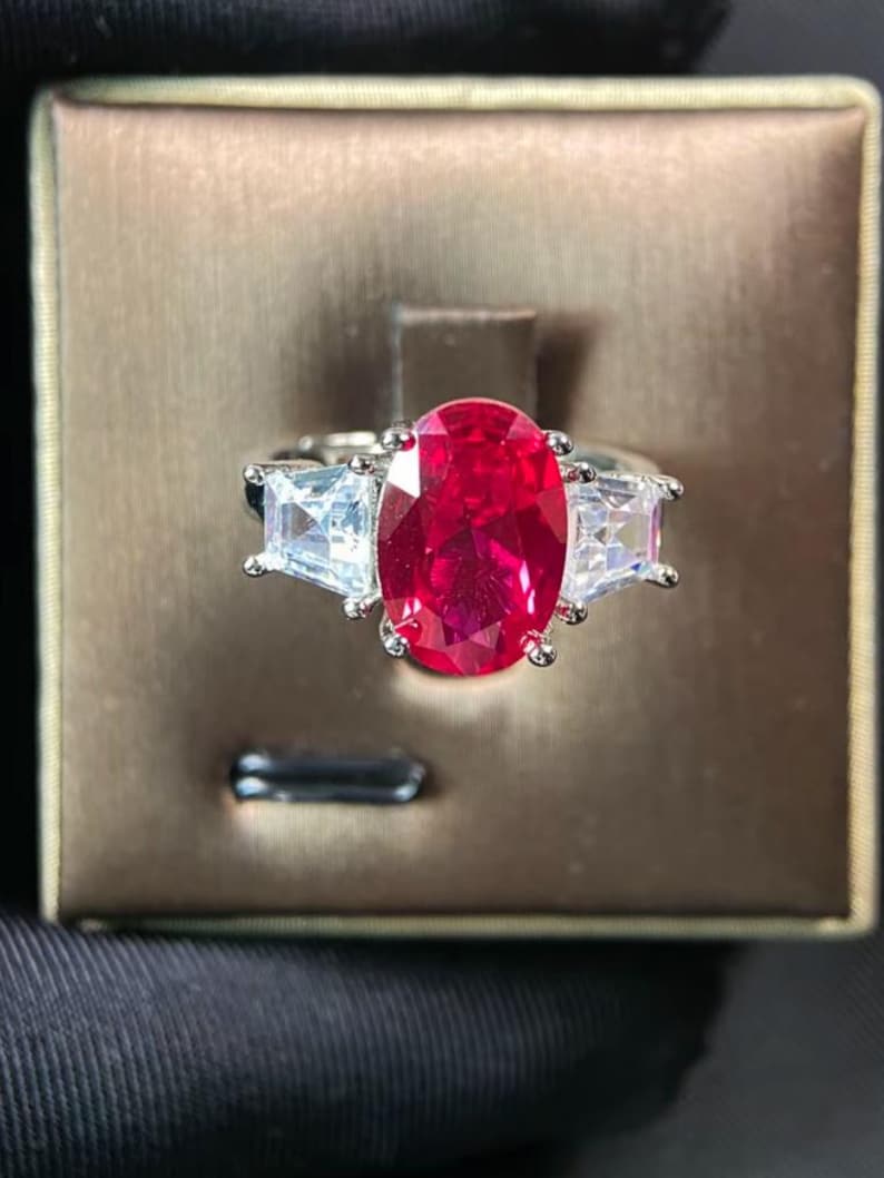 May include: A silver ring with a large, oval-cut, bright red gemstone at its centre, with two smaller, rectangular, clear gemstones either side. The ring is displayed against a brown backdrop.