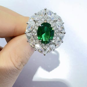 May include: A silver ring featuring a large, oval-cut, emerald-green gemstone at the center, surrounded by smaller, clear, marquise-cut stones. The ring's design is symmetrical, with the smaller stones forming a halo around the central gem.