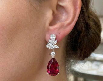 May include: A pair of drop earrings with a cluster of small diamonds at the top, a single diamond in the middle, and a large, pear-shaped, deep red gemstone at the bottom. The earrings are set in silver or white gold.