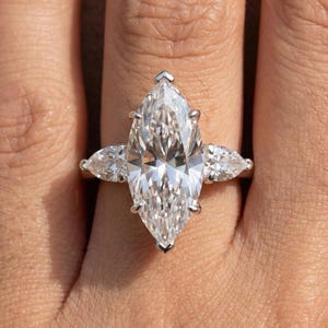 Könnte beinhalten: Ein Verlobungsring mit einem Diamanten im Marquiseschliff und zwei kleineren birnenförmigen Diamanten auf beiden Seiten. Der Ring ist in einem silbernen Band gefasst und wird an einem Finger gezeigt. Der große Mittelstein ist klar und funkelnd.