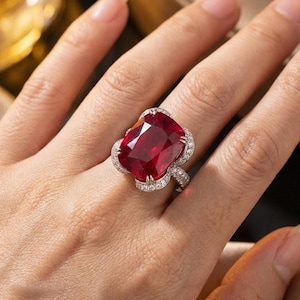 May include: A large, ornate ring featuring a vibrant, cushion-cut red gemstone. The gemstone is surrounded by a halo of smaller, clear stones set in a silver-toned metal band. The ring is displayed on a hand.