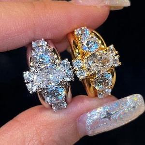 May include: Two diamond rings, one gold and one silver, are held up against a dark background. The gold ring has a large central diamond surrounded by smaller stones. The silver ring has a similar design. Both rings sparkle.