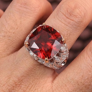 Puede incluir: Un anillo ornamentado con una gran gema roja de corte cuadrado. El anillo tiene una banda plateada con intrincados filigranas y pequeñas piedras claras. Las garras doradas aseguran la gema, creando un contraste llamativo.
