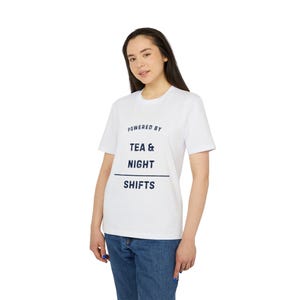 Peut inclure: T-shirt blanc avec l'inscription bleu marine "POWERED BY TEA & NIGHT SHIFTS". Le t-shirt, à col rond, est porté avec un jean bleu. L'ensemble dégage une impression de décontraction.