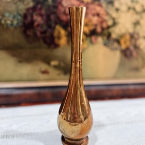 May include: A small, vintage brass vase with a slender, fluted neck and a rounded base. The vase has a warm, golden hue and is displayed on a white lace tablecloth. The background features a blurred painting of flowers.