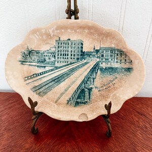 May include: An oval ceramic dish with a scalloped edge and a cream-colored crackle glaze. The center features a dark green transfer print of the Melan Arch Bridge in Waterloo, IA, with buildings and a bridge structure depicted in detail.