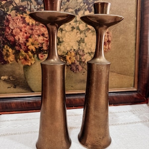 May include: Two bronze-colored candle holders. Each has a flared base, a slender stem, and a wide, flat top with a small cup for holding a candle. The background features a framed floral painting.
