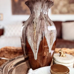 May include: A ceramic vase with a flared rim and a glossy finish, featuring a deep brown base color with white and grey glaze dripping down from the top. The surface has a smooth, reflective texture with vertical ridges along the body of the vase.